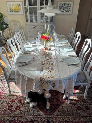 A dining table set for a meal with white chairs, a patterned tablecloth, and a vase of flowers.