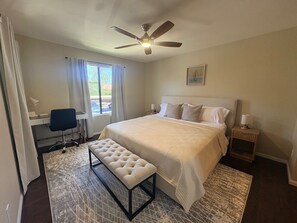 Ein Schlafzimmer mit Bett, Schreibtisch, Stuhl, Bank, Nachttisch, Lampe, Deckenventilator und Fenster mit Vorhängen.