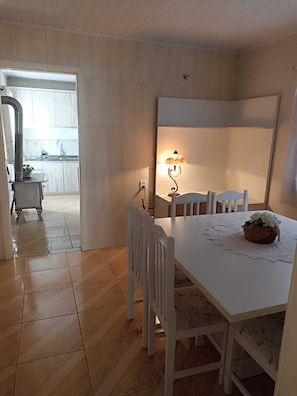 Un coin repas avec une table et des chaises blanches, une lampe fixée au mur et une vue sur une cuisine par une porte ouverte.