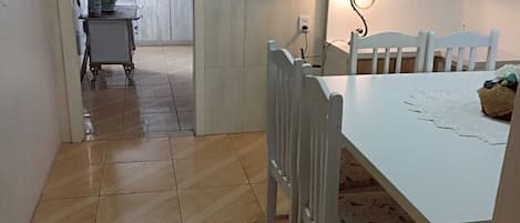 A dining area with a white table and chairs, a small kitchen in the background, and a hallway leading to another room.