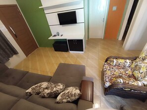 Un salon comprenant un canapé, un fauteuil à motifs, un téléviseur à écran plat fixé sur un meuble blanc et une porte en bois.