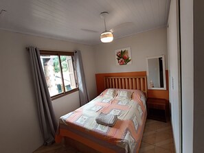 Ein Schlafzimmer mit Bett, einem Fenster mit Vorhängen, einem Deckenventilator und einem wandmontierten Spiegel.