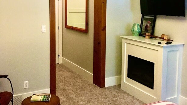 Un dormitorio con cama, televisor sobre la chimenea, un espejo en la pared y una puerta que da a otra habitación.