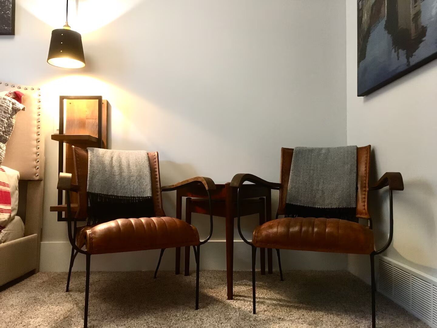 A room with two leather chairs, a small table, a sofa, a hanging lamp, and a framed picture on the wall.