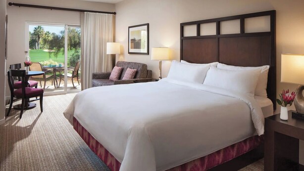 A hotel room with a large bed, a desk with a chair, a sofa, and a view of a golf course through a sliding door.