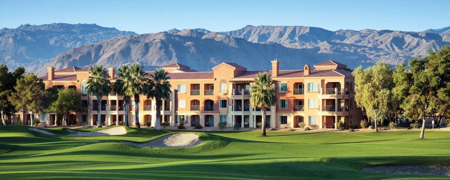 A multi-story hotel with a golf course in front, surrounded by mountains.