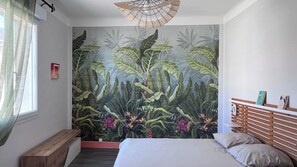 A bedroom with a large mural of tropical foliage, a wooden headboard, a bench, and a bed with pillows. - Vrbo Property (La Tranche-sur-Mer)