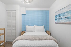 Ein Schlafzimmer mit einem Bett, einem Nachttisch, einem Holztischchen, einer weißen Tür und einem Wandbild.