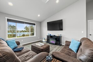 A modern living room with leather sofas, a fireplace, and a large window overlooking a body of water. - Cozy Cove - Hot Tub, Waterfront, Game Room Sleeps 6 (Ocean Shores)