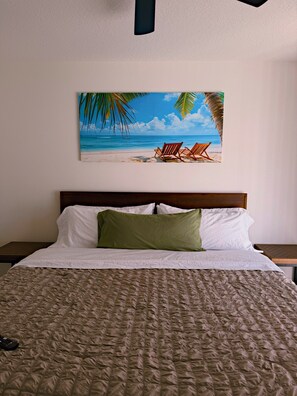 A hotel room with a bed, a painting of a beach scene, and a nightstand with a lamp. - The Sailfish (Stuart)