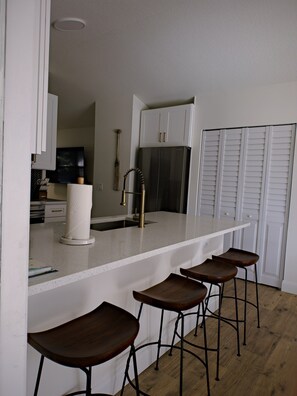 A kitchen with a white countertop, stainless steel refrigerator, and wooden stools. - The Sailfish (Stuart)