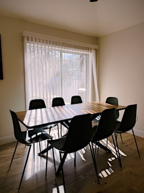A dining area with a wooden table and black chairs, a window with white curtains, and a ceiling fan. - The Sailfish (Stuart)