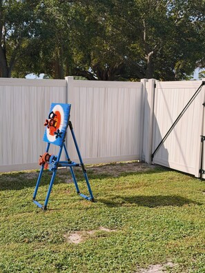 A blue easel with a painting in a grassy area, a white fence, and trees in the background. - The Sailfish (Stuart)