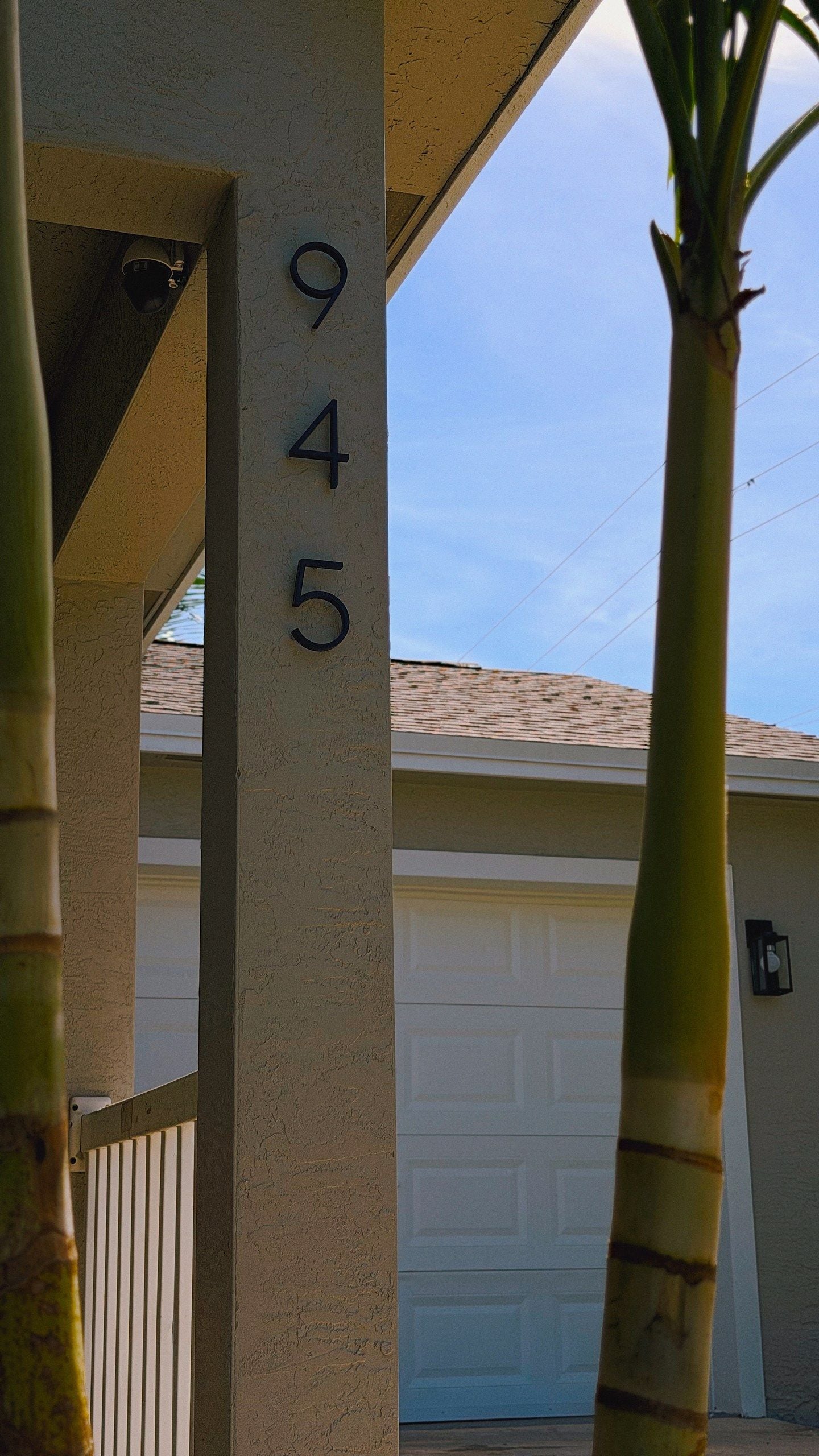 A house number 945 on a beige wall, with a garage door and a clear sky in the background.