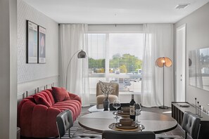 A modern living room with a red sofa, a dining table set for two, and a large window with blinds. - Casa Positano® Designer 2BR/2BA in Irvine w/ Pool (Irvine)