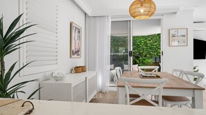 A modern dining area with a wooden table, white chairs, a white console, and a large potted plant. - Beach Lane | 250m to the beach | Accom Holidays (TERRIGAL)