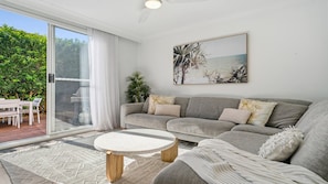 A living room with a grey sofa, a round coffee table, and a large window with a view of greenery. - Beach Lane | 250m to the beach | Accom Holidays (TERRIGAL)