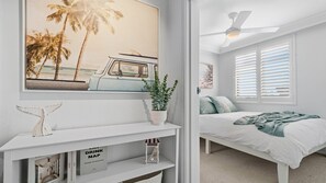 A bedroom with a bed, a ceiling fan, a window with blinds, a wall-mounted TV, and a shelf with decorative items. - Beach Lane | 250m to the beach | Accom Holidays (TERRIGAL)