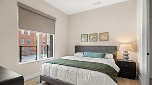 Quarto com cama, mesa de cabeceira, luminária e janela com persianas.