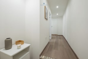 Un couloir avec un mur blanc, un plancher en bois, une porte blanche au fond et une petite table sur laquelle se trouvent un vase et une bougie.