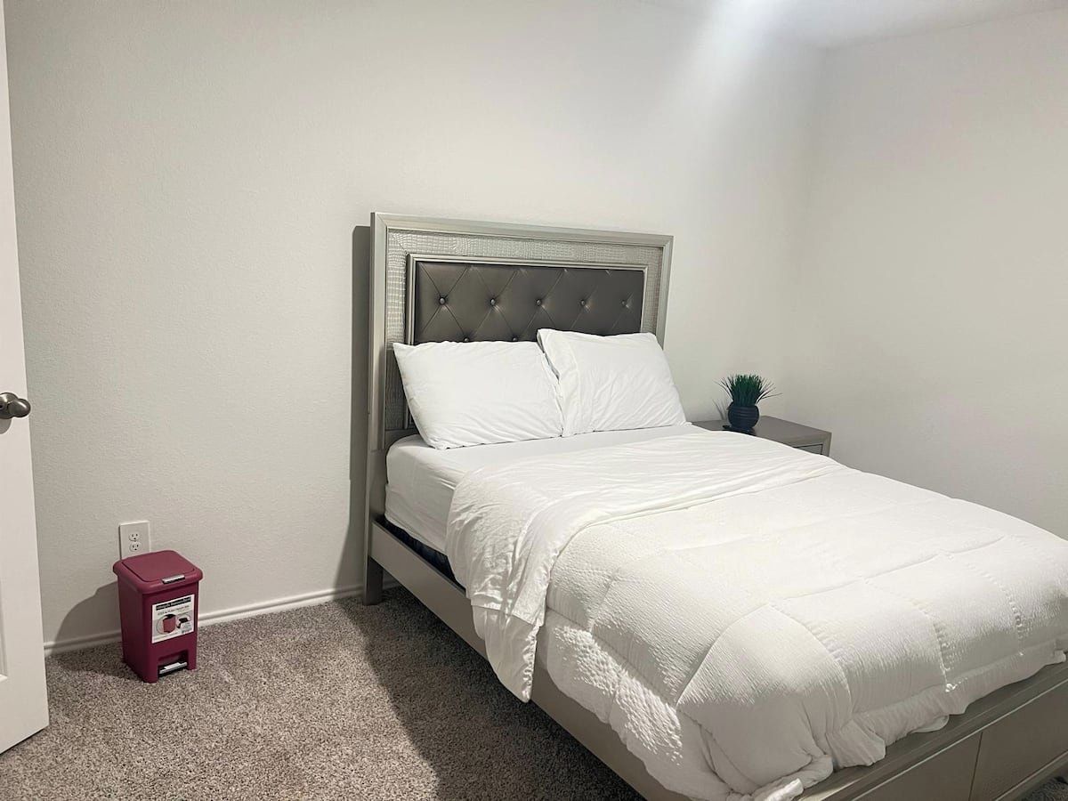 A single bed with a tufted headboard, white bedding, and a small red trash bin beside the wall.