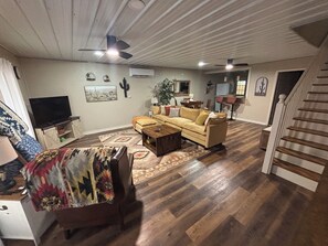 Un salón amplio con sofá, mesa de centro y televisor. Se ve una escalera que lleva a otro nivel.