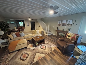 Sala de estar com sofá, poltrona, mesa de centro e tapetes estampados. Uma escada com corrimão é visível ao fundo.
