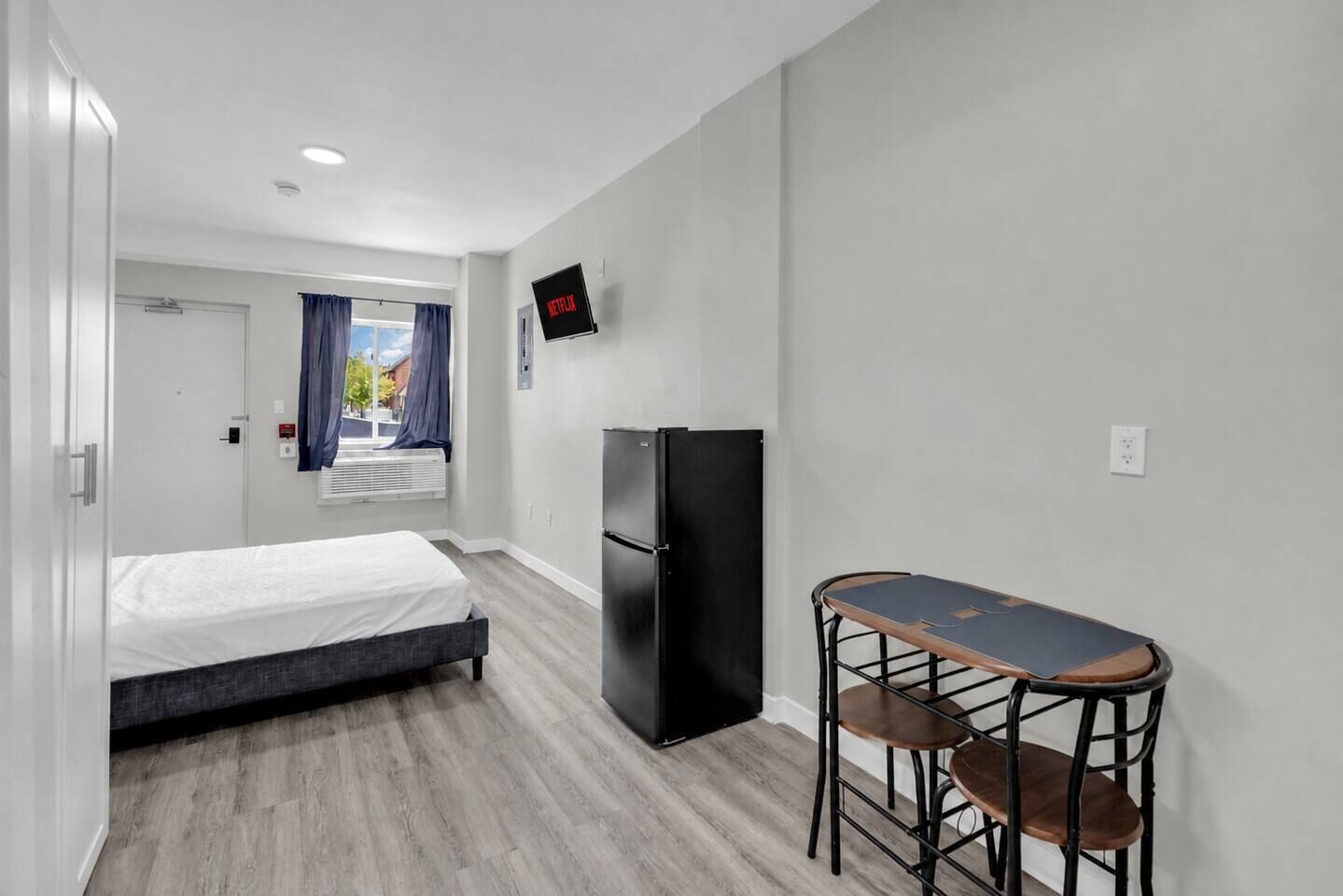 Ein kleines Hotelzimmer mit Bett, Kühlschrank, Fernseher und einer kleinen Essecke.