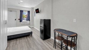 Quarto de hotel compacto com cama, geladeira, TV e pequena área de refeições.