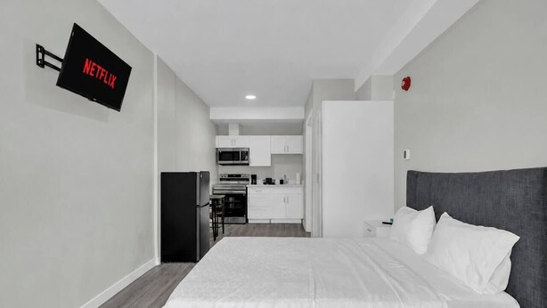 Quarto de hotel moderno com cama, TV de tela plana na parede, cozinha compacta e banheiro privativo.