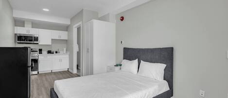 A modern bedroom with a large bed, a built-in wardrobe, and a kitchenette in the background.