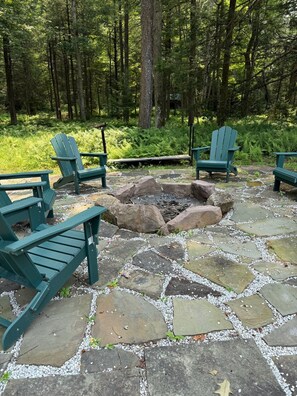 A stone fire pit surrounded by Adirondack chairs in a forested area.