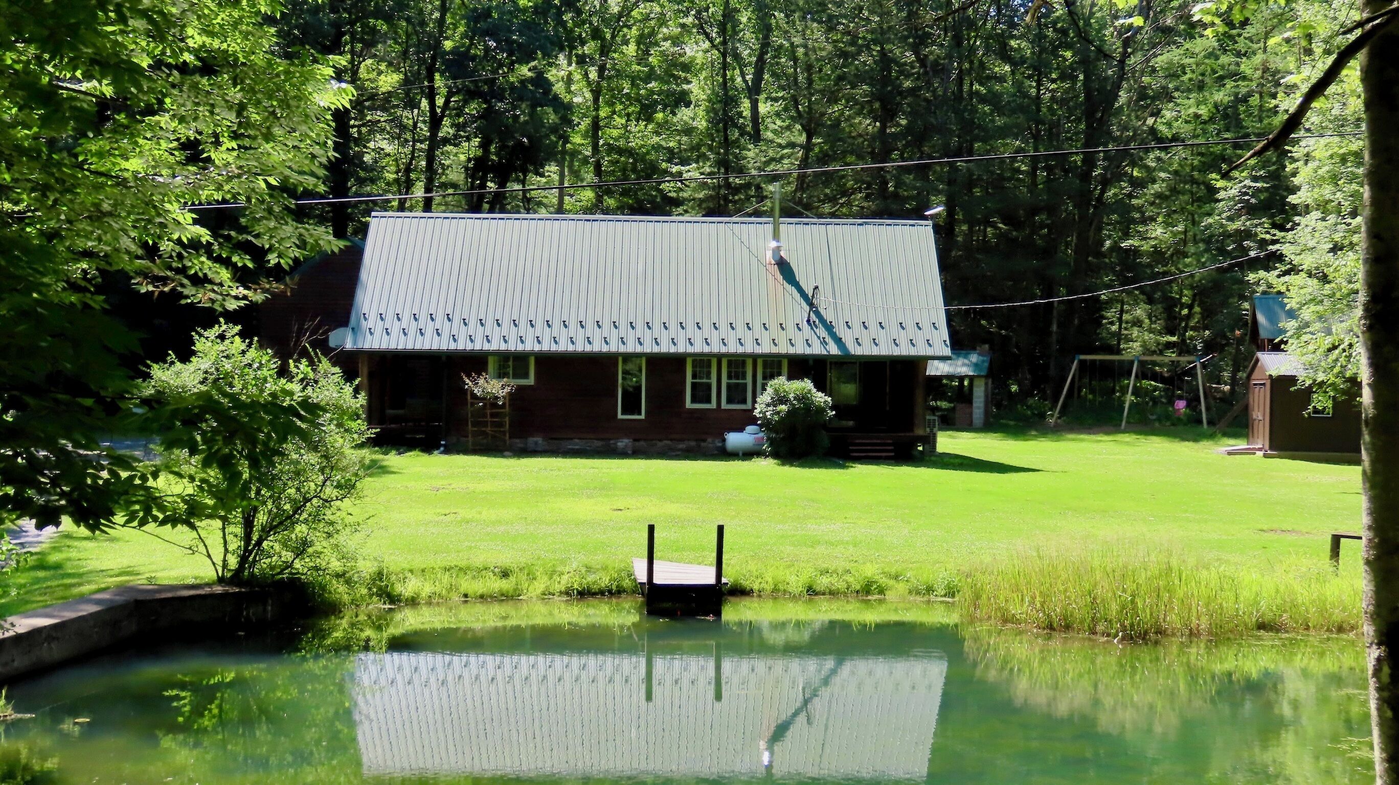 Une cabane avec un toit en métal, entourée d’une cour verdoyante et d’un étang.