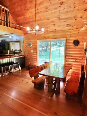 A cabin interior with a dining table, benches, a kitchen area, and a large window.
