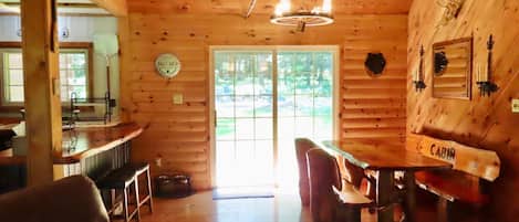 A cabin interior with a wooden ceiling, a dining table with chairs, a bar area, and a large window.