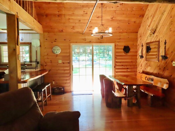 A cabin interior with a wooden ceiling, a dining table with chairs, a bar area, and a large window.