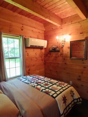 A wooden cabin room with a bed, a window, an air vent, a wall-mounted light fixture, and a framed picture.