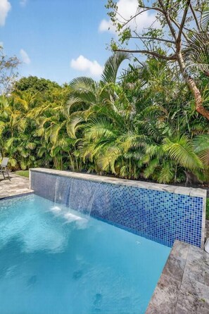 A swimming pool with a tiled edge, surrounded by lush greenery and a clear sky. - Pink Palm Retreat - Pineapple Grove (Delray Beach)