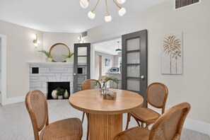 A dining area with a round wooden table and wicker chairs, a fireplace with a stone facade, and a door leading to another room. - Pink Palm Retreat - Pineapple Grove (Delray Beach)