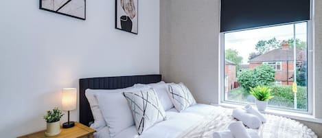 A bedroom with a bed, bedside table, lamp, and window with a view of houses.
