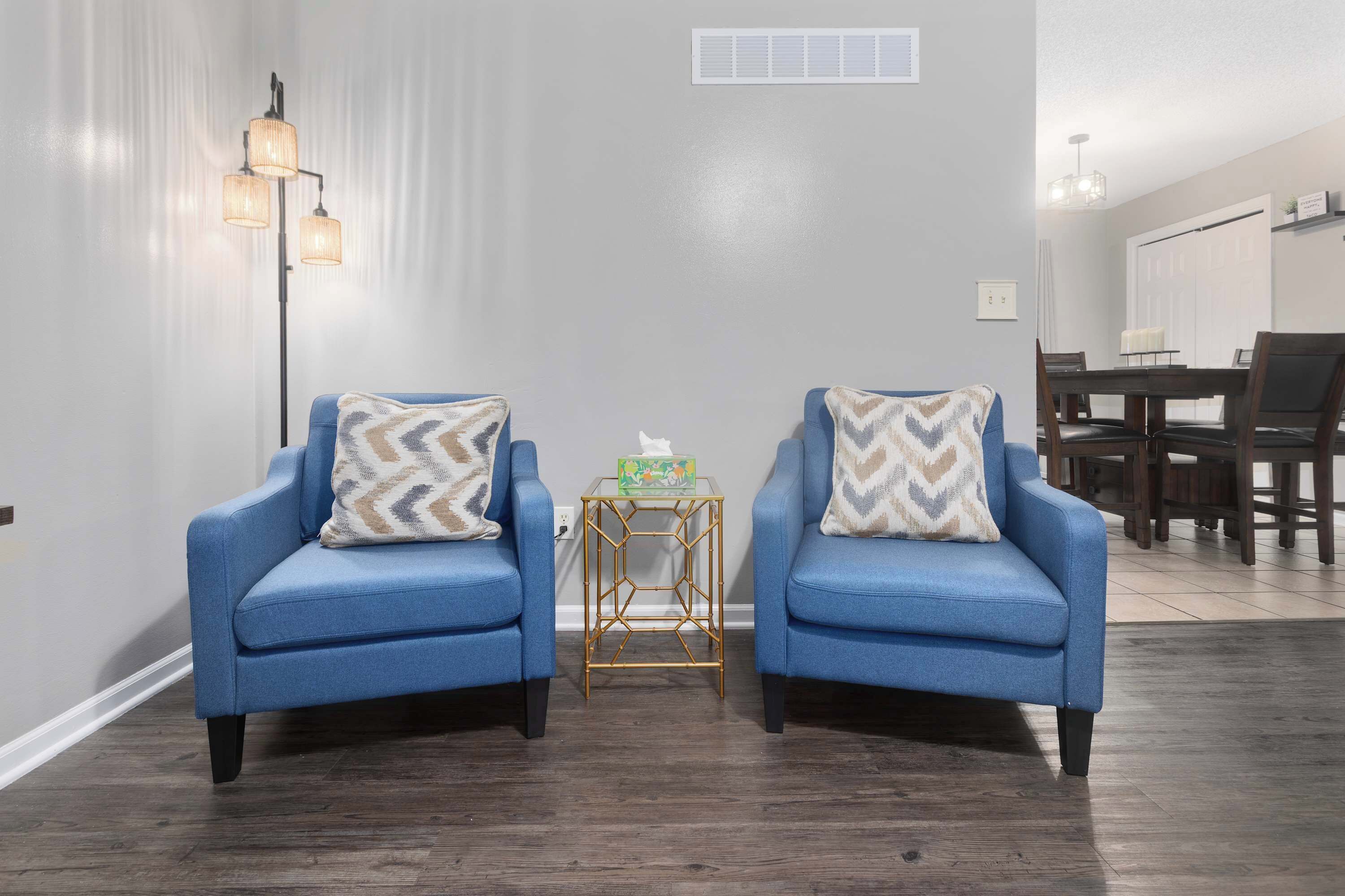 A room with two blue armchairs, a small side table, and a dining area in the background.