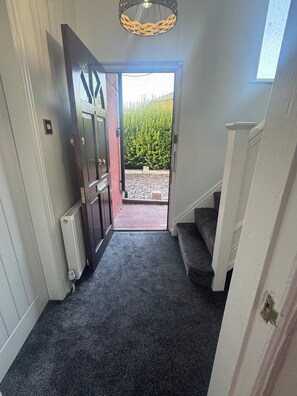 Un couloir avec un sol recouvert de moquette, menant à une porte, un radiateur et un escalier.