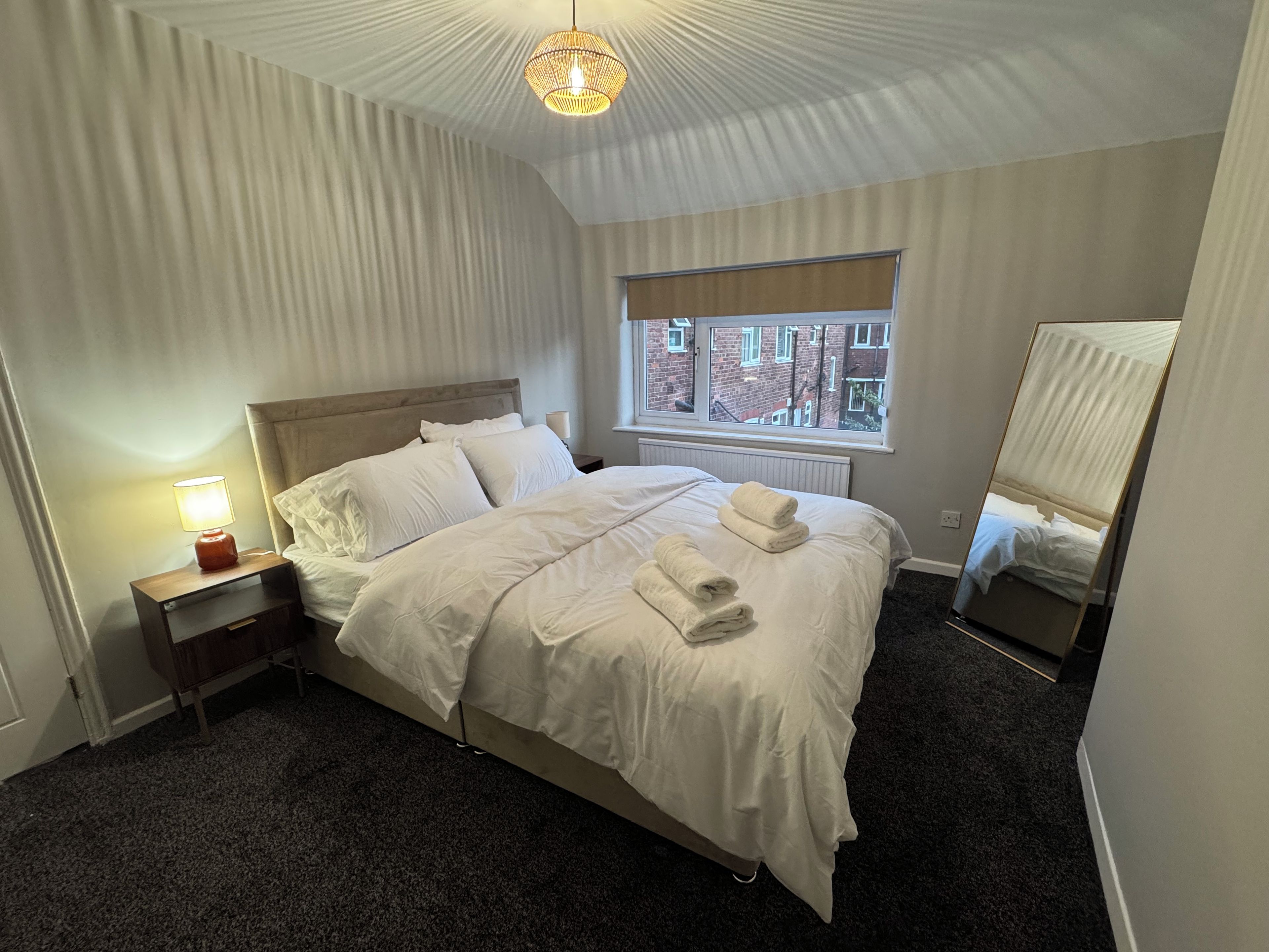 A neatly made bed with white linens and towels, a bedside lamp, a window with a view of buildings, and a ceiling light fixture.