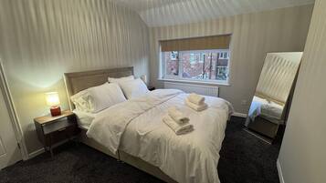 A neatly made bed with white linens and towels, a bedside lamp, a window with a view of buildings, and a ceiling light fixture.