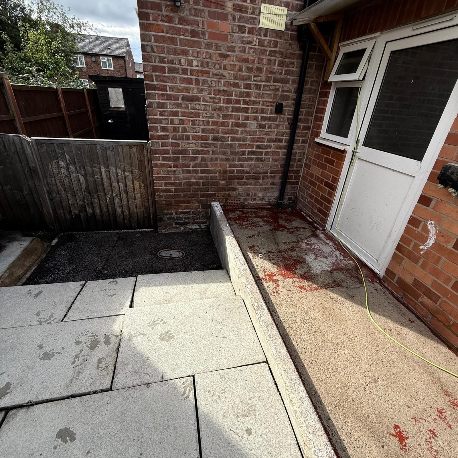 A brick wall with a window and a door, a concrete floor with a drain, and a wooden fence.