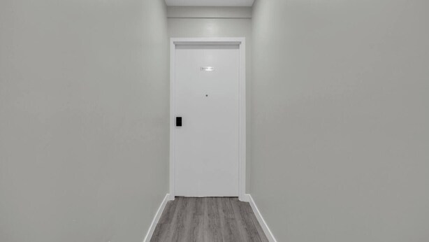 A narrow hallway with a white door at the end, a light switch on the wall, and a wooden floor.