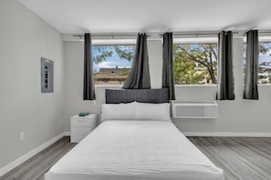 A modern bedroom with a large bed, a nightstand, a window with curtains, and an air conditioning unit. - The Premier Suite • Studio • DT Oshawa (Oshawa)