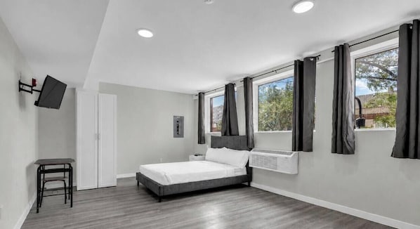 A bedroom with a bed, a desk, a chair, a TV mounted on the wall, and a window with curtains. - The Premier Suite • Studio • DT Oshawa (Oshawa)