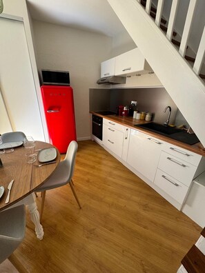 A compact kitchen with white cabinets, a red refrigerator, and a dining area with a wooden table and chairs. - ExVoto - Charming Apartment in the Heart of Pézenas (Pézenas)