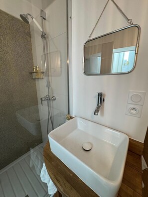 Een badkamer met een douche in een glazen cabine, een wit wastafelmeubel met zeepbakje en een spiegel met een ketting.
